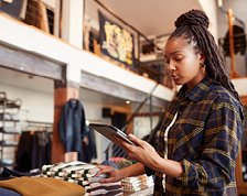 A store manager in a plaid shirt stands in a retail store and reads from the tablet in her hand. 